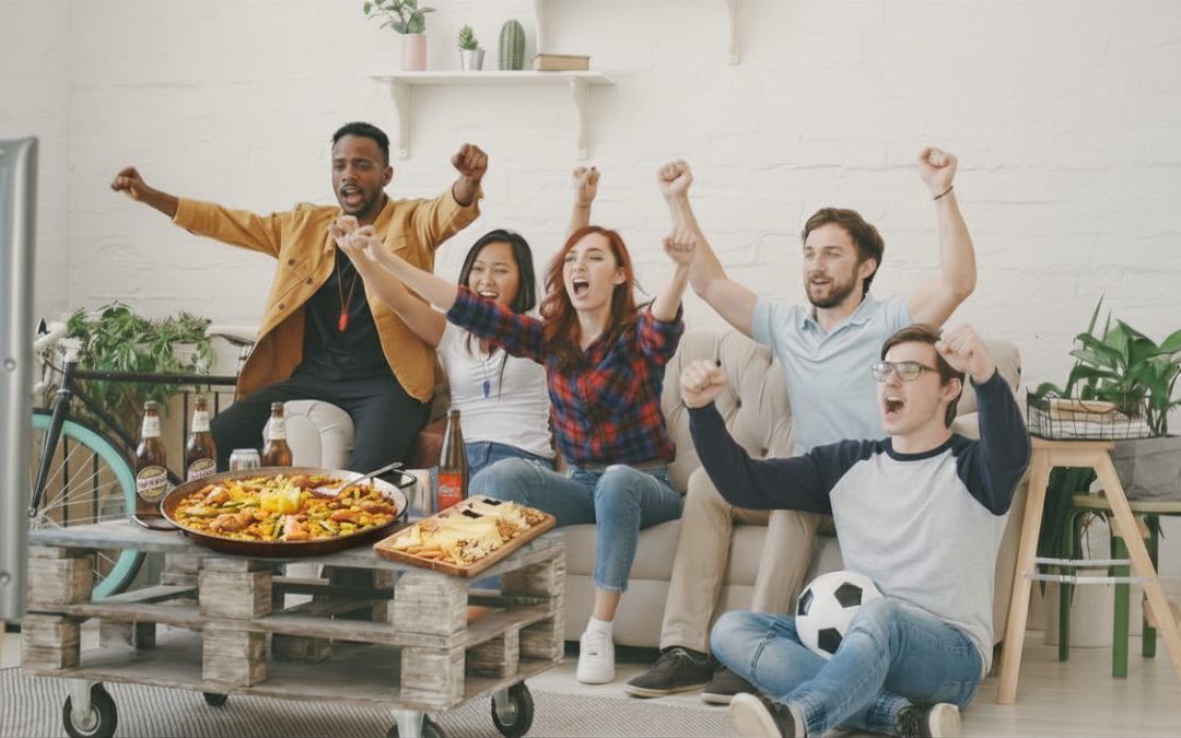 Grupo de amigos compartiendo raciones y una paella para llevar frente al televisor viendo el fútbol